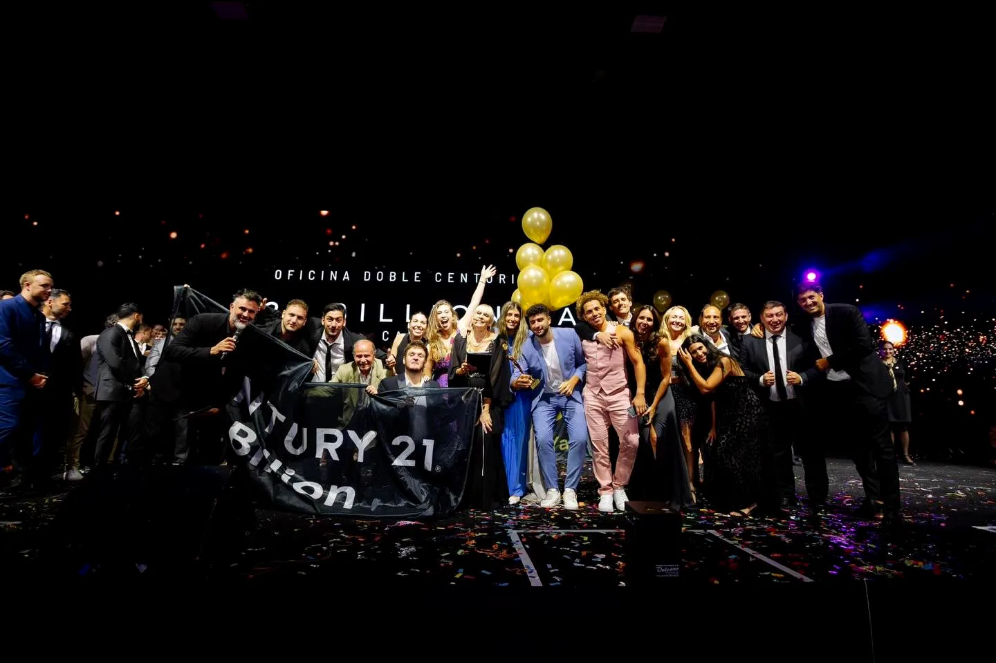 El equipo de Century 21 Billion en el escenario de la Convención Century 21, con la bandera de la oficina, globos dorados y papelitos